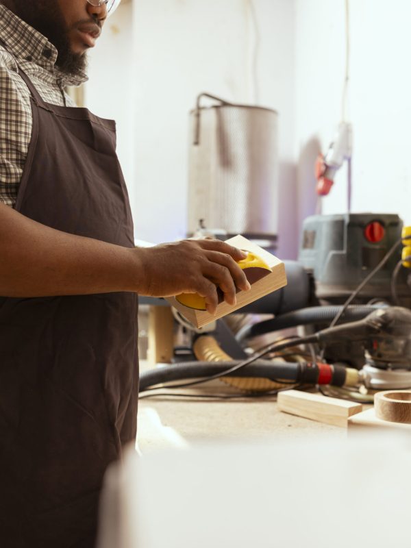 African american man in assembly shop using sandpaper for sanding wooden surface before painting it, ensuring adequate finish. BIPOC carpenter using abrasive sponge to repair damages on wood block