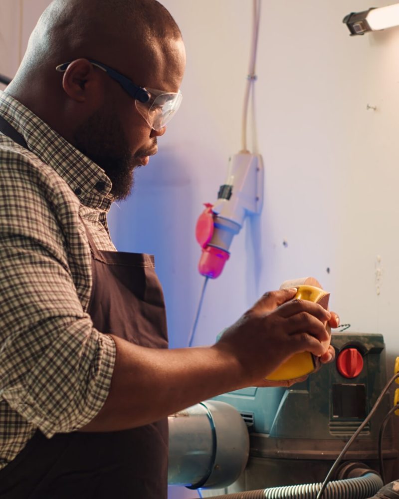 BIPOC man in studio with woodworking tools in background using sandpaper for sanding wooden surface. Woodworking expert using abrasive sponge to fix damages suffered by piece of wood, camera A
